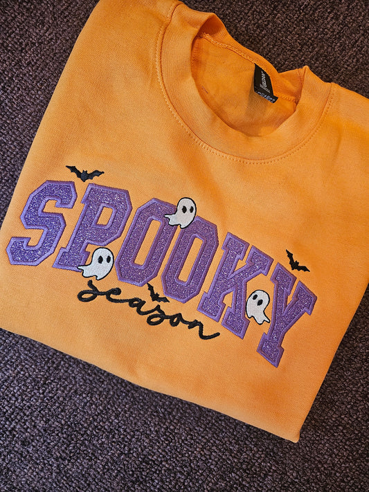 Orange crewneck sweatshirt laid flat, with purple glitter SPOOKY applique lettering, the word season in black cursive embroidery, and small white ghosts and black bats