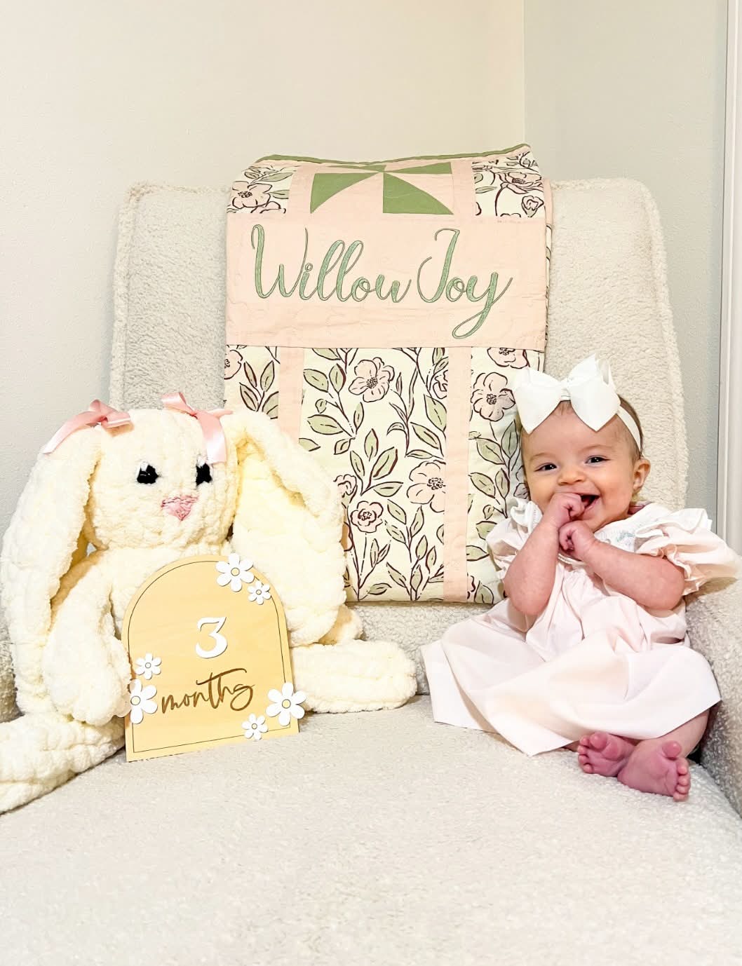 A baby girl in a pale pink dress and white bow sits on a cream armchair next to a large cream stuffed bunny and a floral quilted cushion with the words 'Willow Joy' and a yellow sign reading '3 months'.