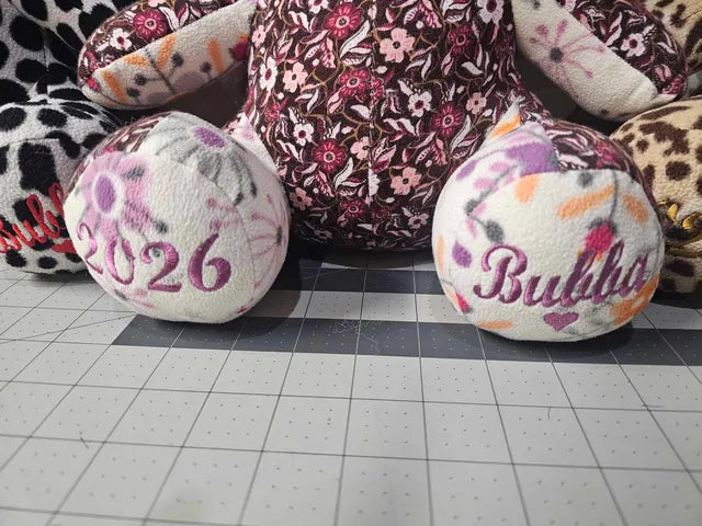A handmade plush toy or cushion with a floral patterned body and two round white feet embroidered with '2026' and 'Bubba', displayed on a cutting mat.