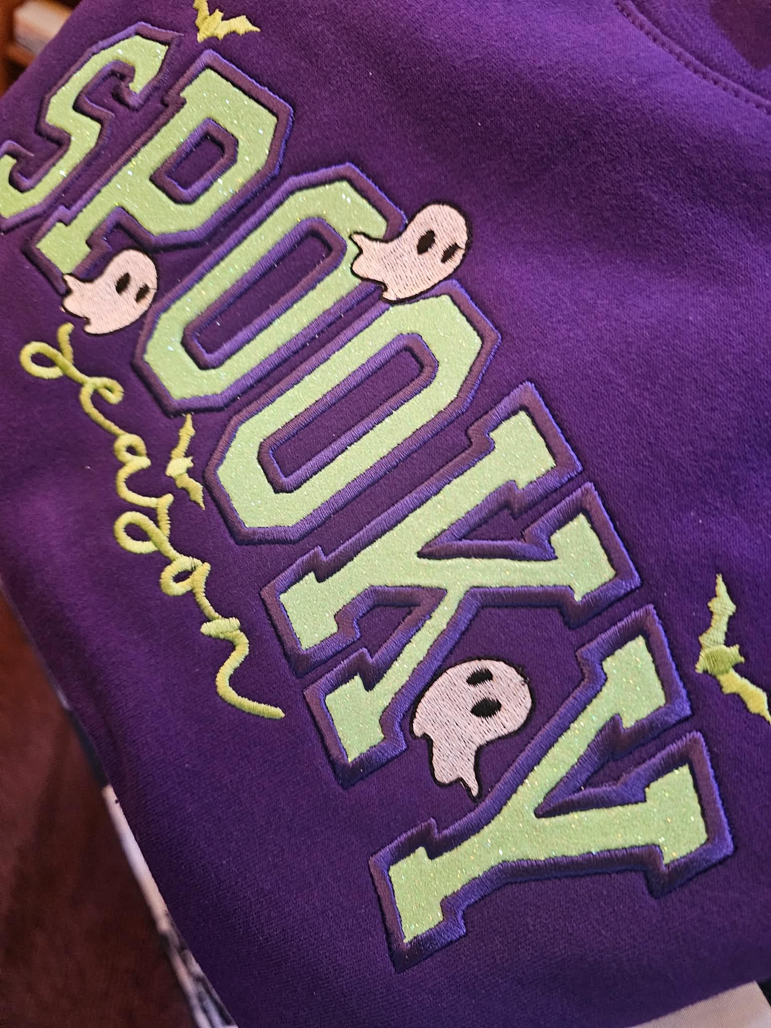 Close-up of green glitter Spooky Season applique on a purple crewneck, with the word season in cursive embroidery, two stitched ghosts, and small bats