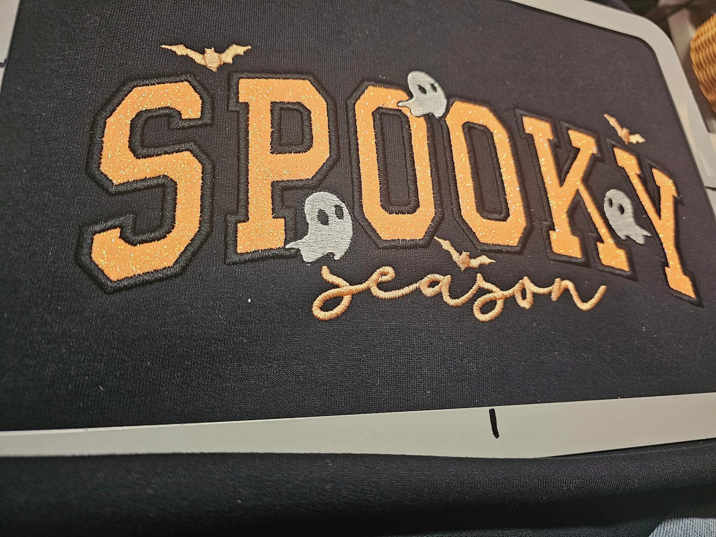 Close-up of neon orange glitter Spooky Season applique on a black crewneck, with the word season in tan cursive embroidery, gray stitched ghosts, and small bats