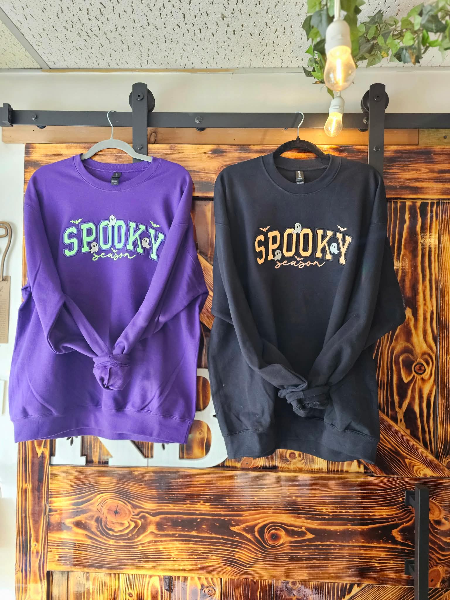 Two finished Spooky Season crewnecks hanging in the Krafty Bandit shop in Belle Vernon, PA: a purple sweatshirt with green glitter applique lettering and a black sweatshirt with neon orange glitter applique lettering