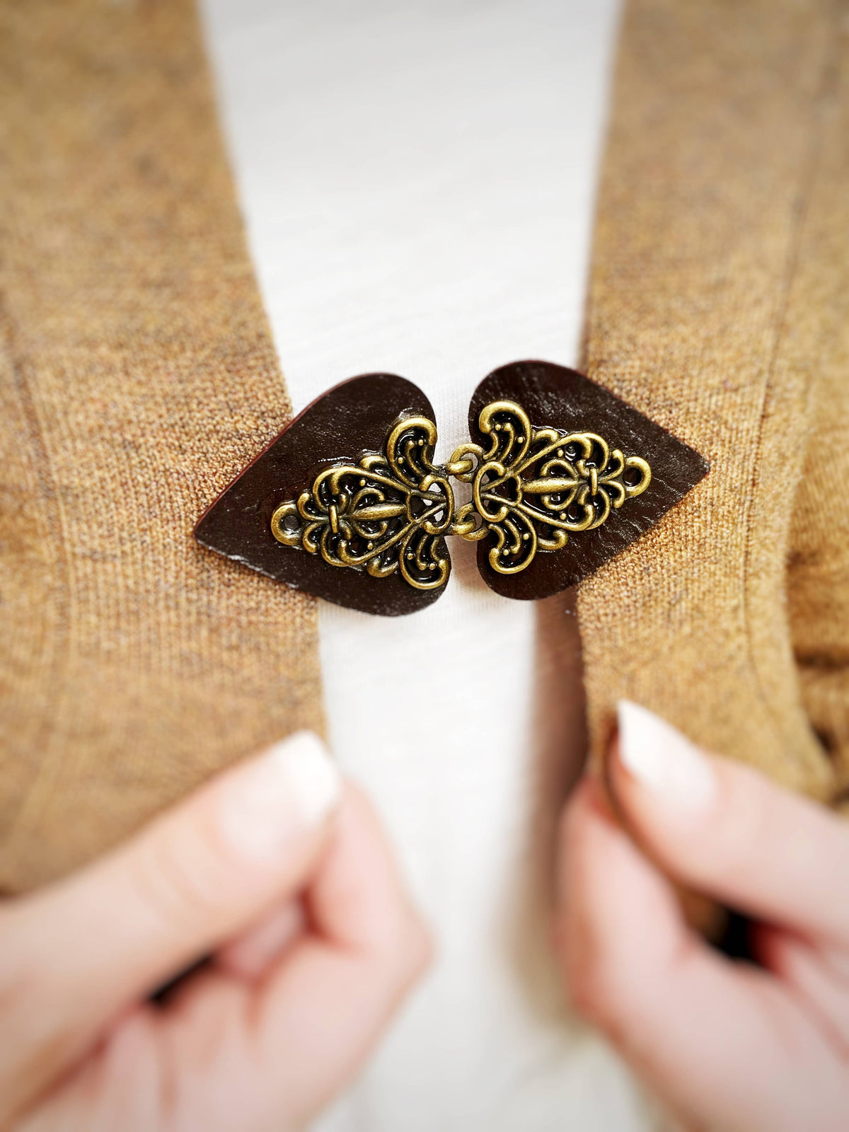 Hands fastening the dark brown filigree clasp on a camel cardigan