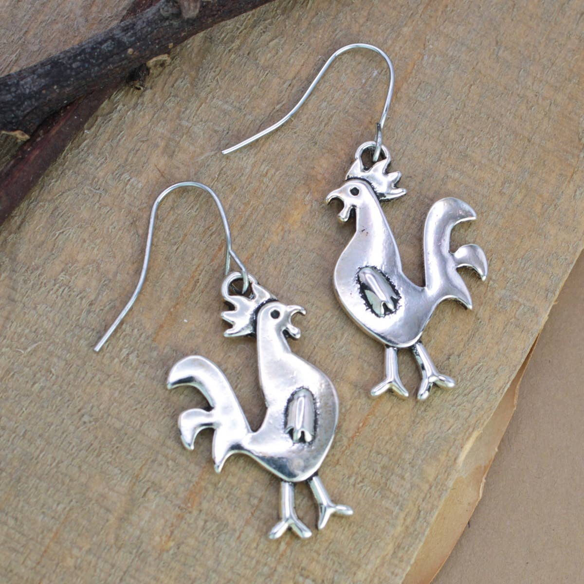 Pair of silver tone chicken earrings with combs and tail feathers on a wooden surface