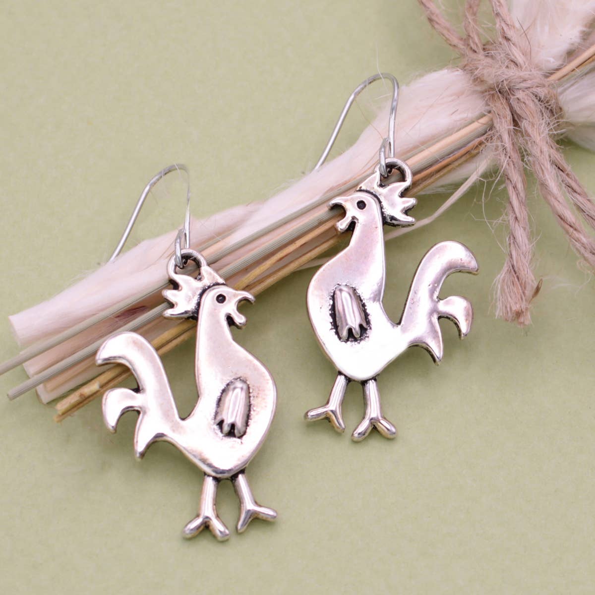 Pair of silver tone chicken earrings displayed on pink twine