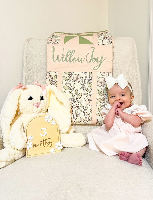 A baby girl in a pale pink dress and white bow sits on a cream armchair next to a large cream stuffed bunny and a floral quilted cushion with the words 'Willow Joy' and a yellow sign reading '3 months'.