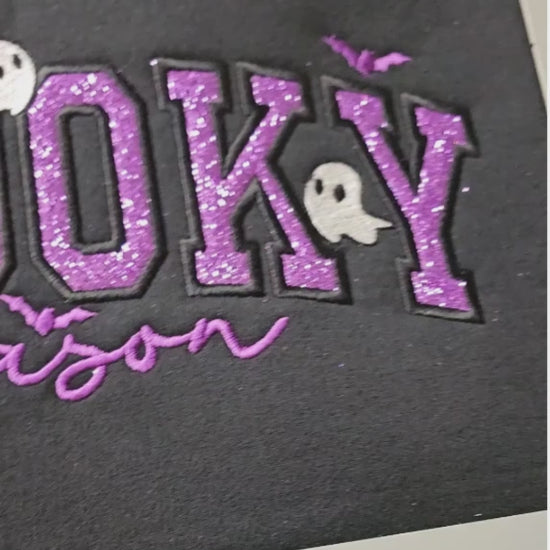 Close-up video of the Spooky Season applique embroidery on a black crewneck, showing purple glitter letters outlined in dense black stitching, a small white ghost, and bat accents