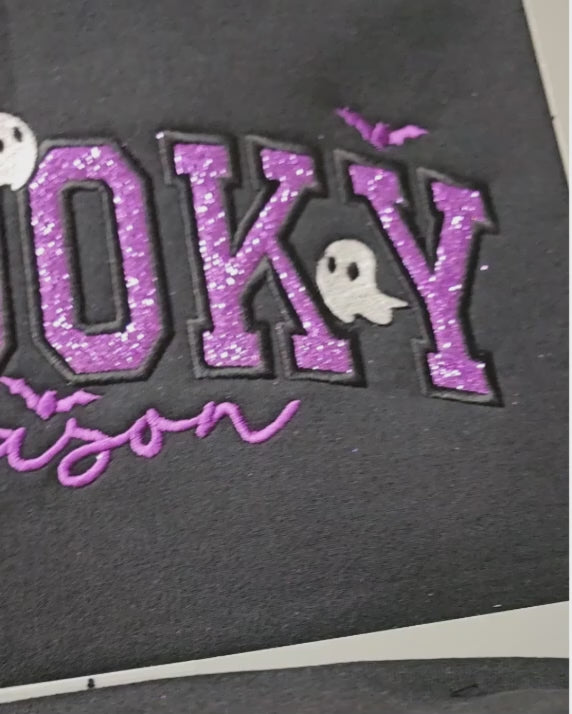 Close-up video of the Spooky Season applique embroidery on a black crewneck, showing purple glitter letters outlined in dense black stitching, a small white ghost, and bat accents