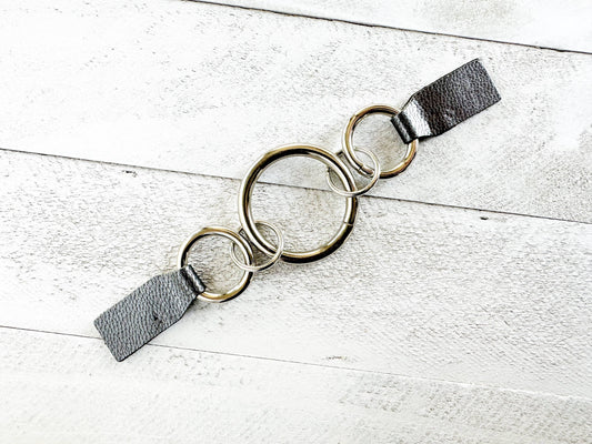 Three interlocking silver rings with gray leather grip tabs, flat on white wood