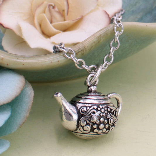 Silver tone floral etched teapot pendant necklace on a chain, displayed with a flower